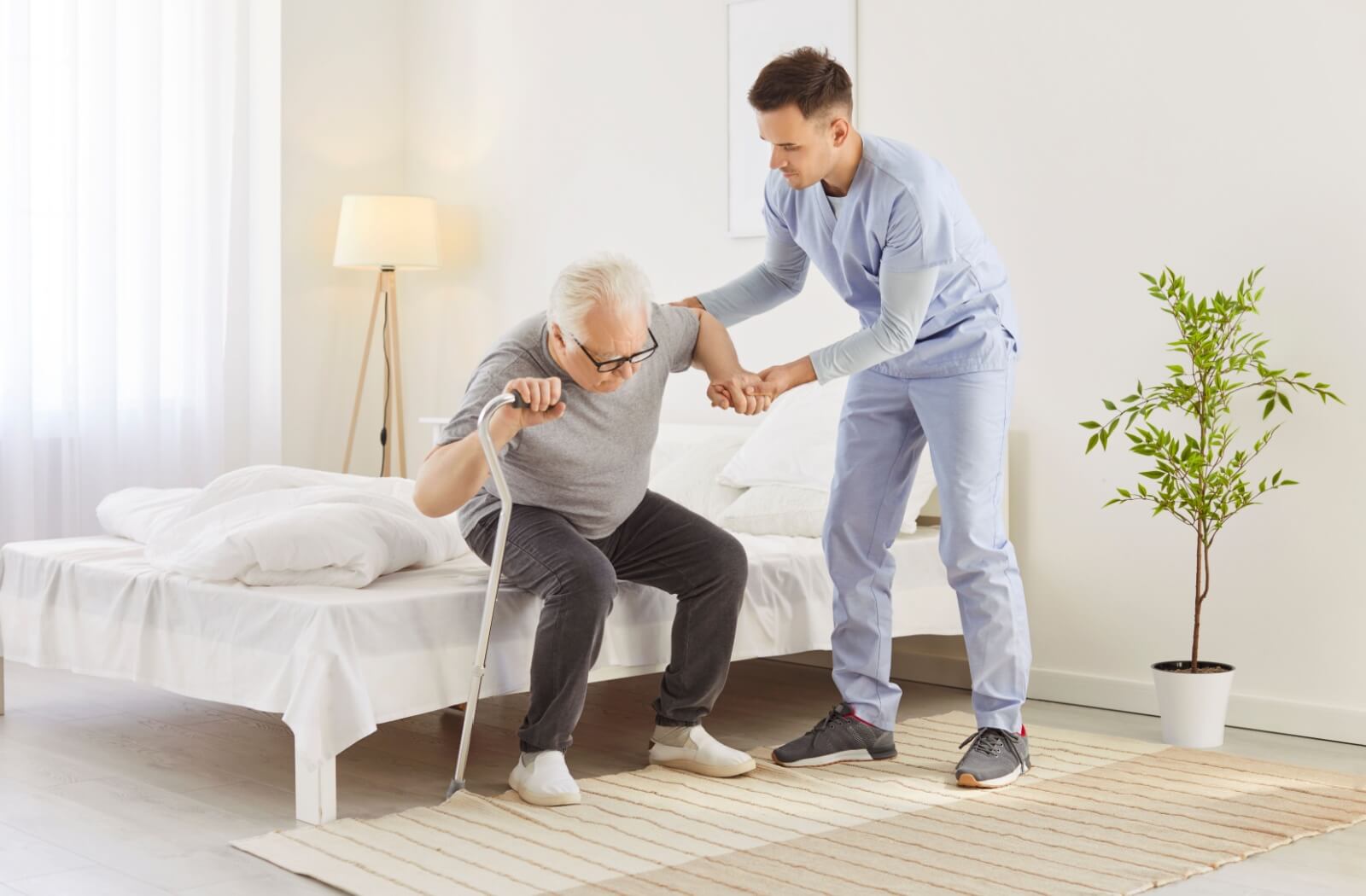 A caregiver helping a senior man with a cane stand up from a bed.